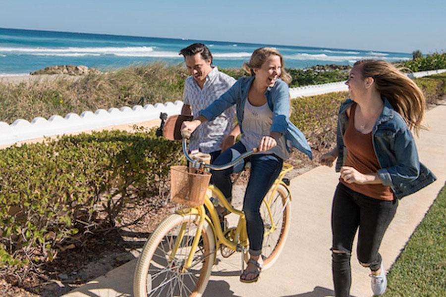 Three PBA students, one holding a skateboard, one riding a bike, and one walking, on a path near the beach.