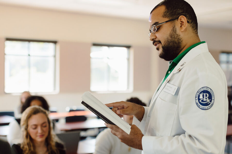 a male student standing in a classroom reading from a textbook