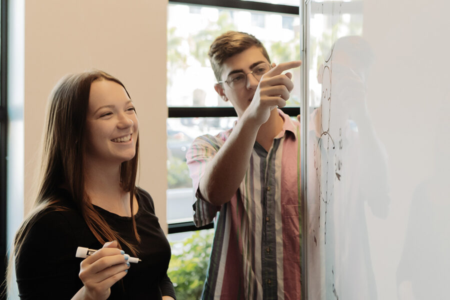 Two PBA students draw diagrams on a whiteboard.