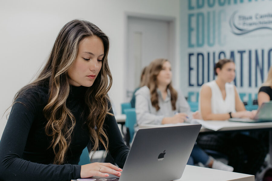 Students take notes on laptops during a lecture in a classroom.